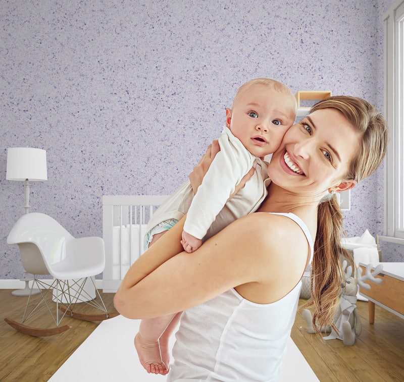 Eine Mutter hält ihr Baby und lächelt. Sie sind in einem Babyzimmer, dessen Wände mit Belka Tapete Flieder verkleidet sind.