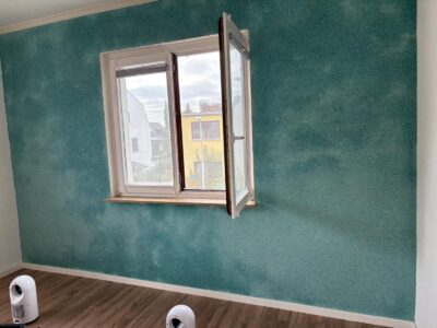 Eine teilweise fertiggestellte Wand mit der B39 Türkis Tapete, die ihre lebendige türkise Textur zeigt. Eine Leiter steht in der Nähe, und ein Fenster lässt natürliches Licht in den Raum.