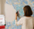 Eine Person trägt Belka Tapete mit einer Kelle auf eine Wand mit blau-goldenem Muster, während ein moderner Raum mit Sofa und Tisch im Hintergrund sichtbar ist.
