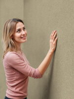 Beige Belka Y2503 Tapete Leinenbeige, aufgetragen auf eine Wohnzimmerwand in moderner Optik. Die natürliche, strukturierte Oberfläche harmoniert mit Holzmöbeln und weißer Deko und schafft eine schlichte, elegante Atmosphäre. Die Tapete zeigt im Licht einen warmen, leicht glänzenden Ton.