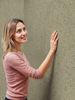 Wüstentaupenfarbene Belka Y2504 Tapete, aufgetragen auf eine moderne Wohnzimmerwand. Die warme, erdige Textur harmoniert mit minimalistischen Möbeln in Grau und Weiß, schafft eine gemütliche, stilvolle Atmosphäre. Im Licht schimmert die Tapete sanft und betont die natürliche Eleganz des Raumes