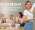 Junge Frau trägt Belka Tapete an die Wand auf, während Mutter und Tochter im gemütlichen Wohnzimmer sitzen – DIY-Anwendung mit natürlichen Materialien.