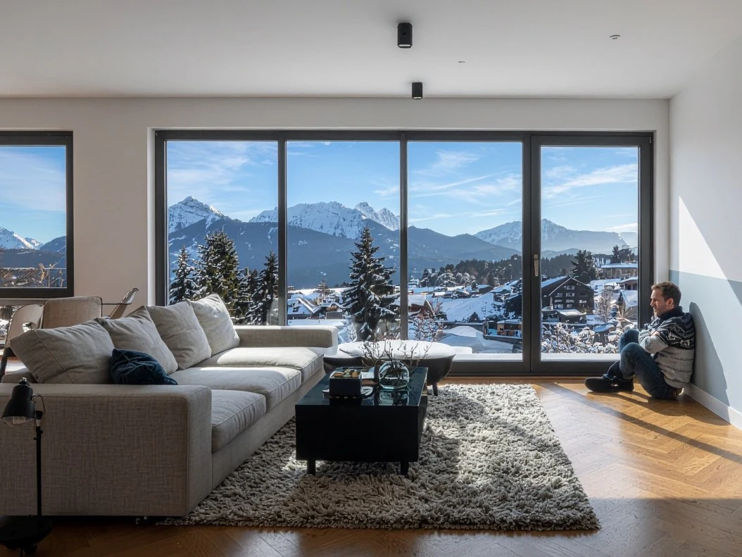 Mann sitzt frierend an kalter Wohnzimmerwand mit großem Fenster und verschneiter Berglandschaft im Hintergrund